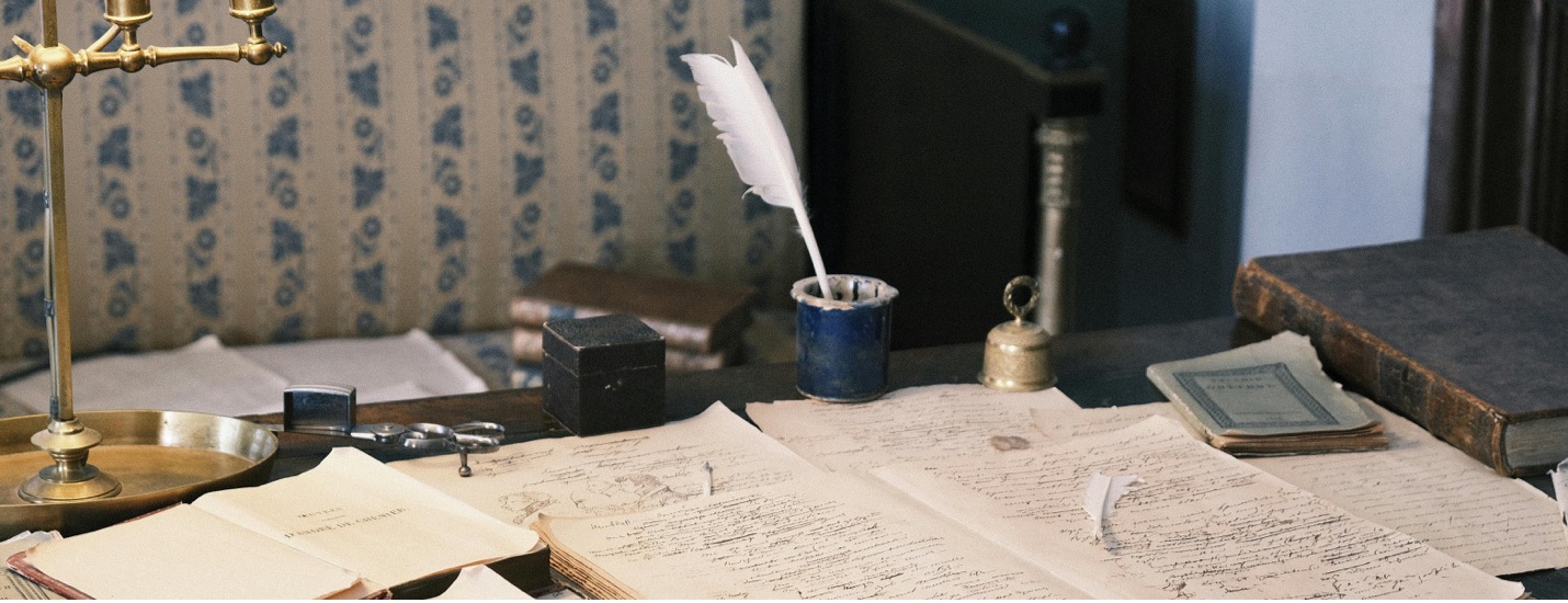 Antique desk with ledgers spread out, books, paper weights, and a plume pen.